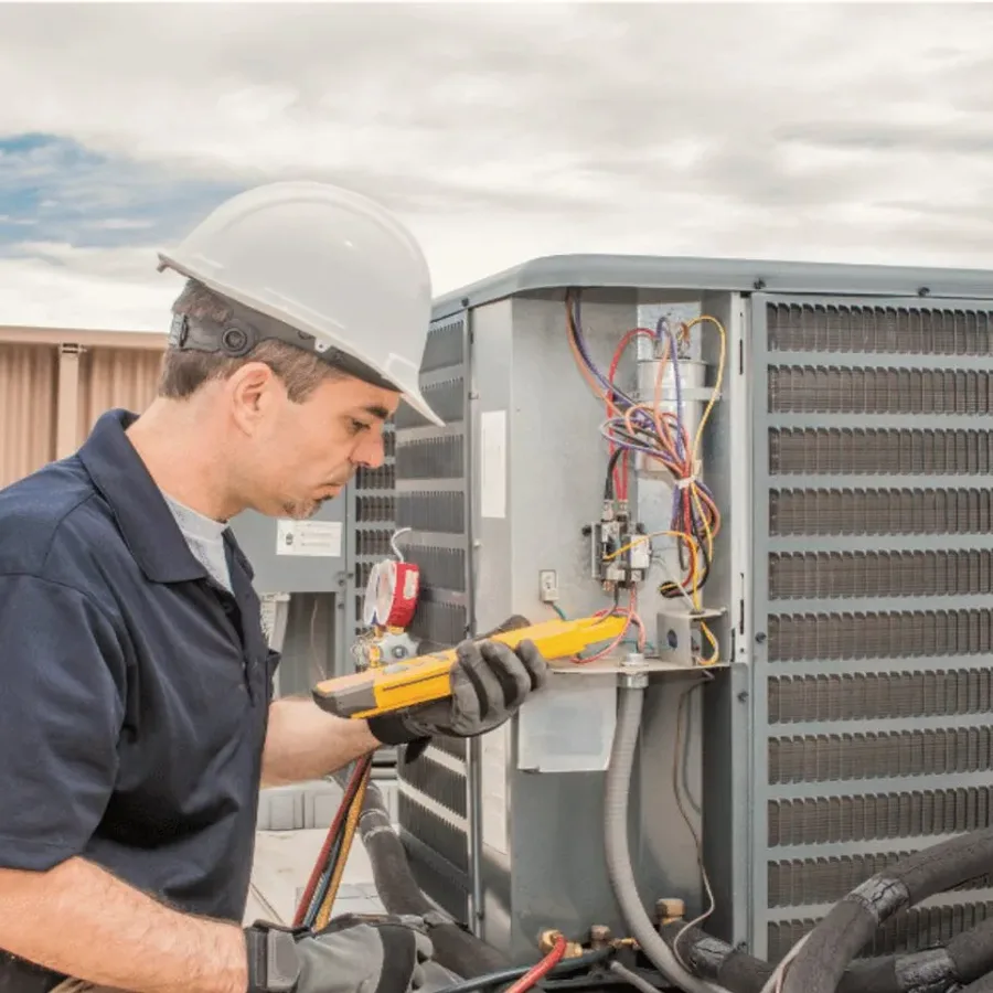 HVAC technician in a hard hat testing an outdoor air conditioner with a multimeter